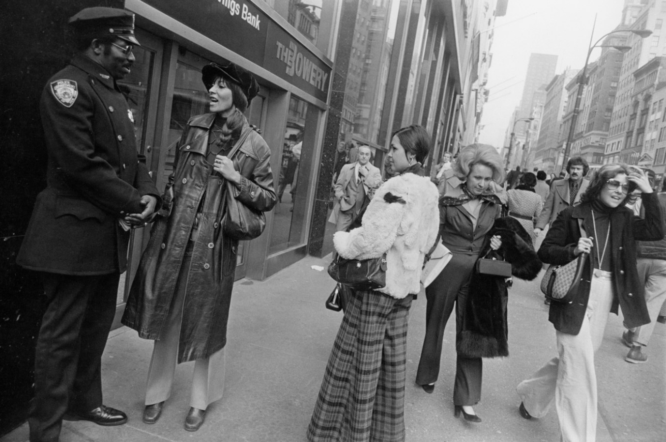 Black-and-white photograph of two young women pausing on a busy city sidewalk to talk to a police officer