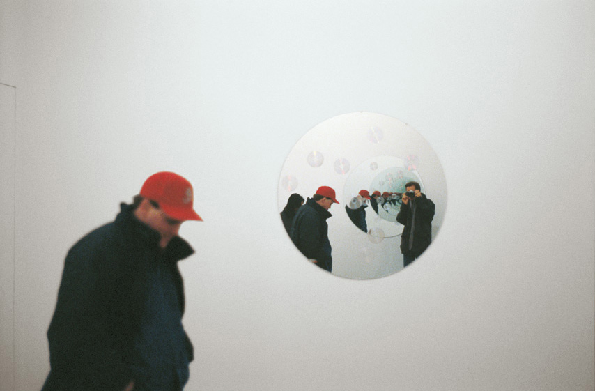 An installation shot of a person in front of a circular mirror on a white wall reflecting him and the mirror behind him