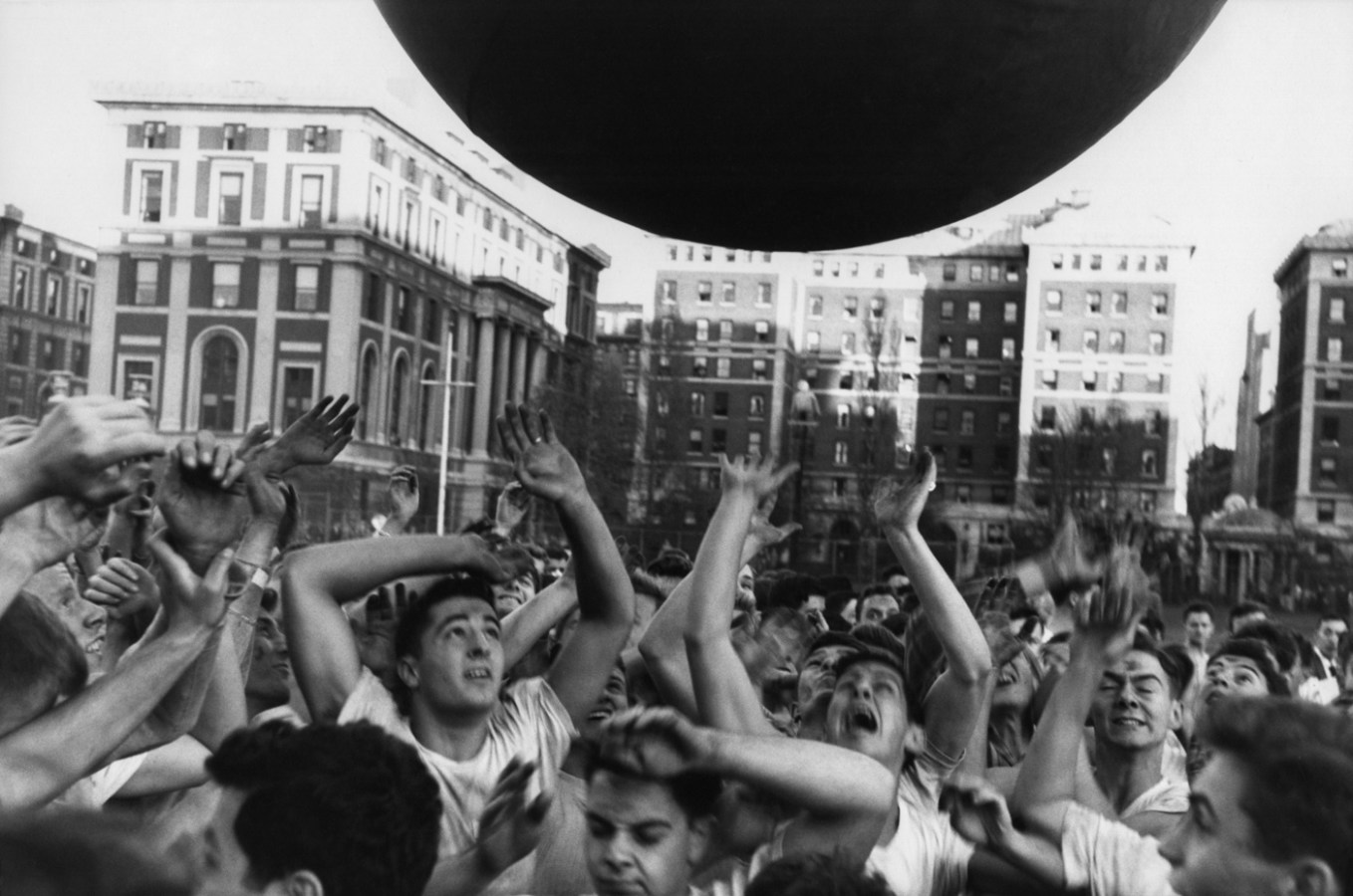 Black-and-white photograph of a crowd of young men with arms raised towards a round object floating above them