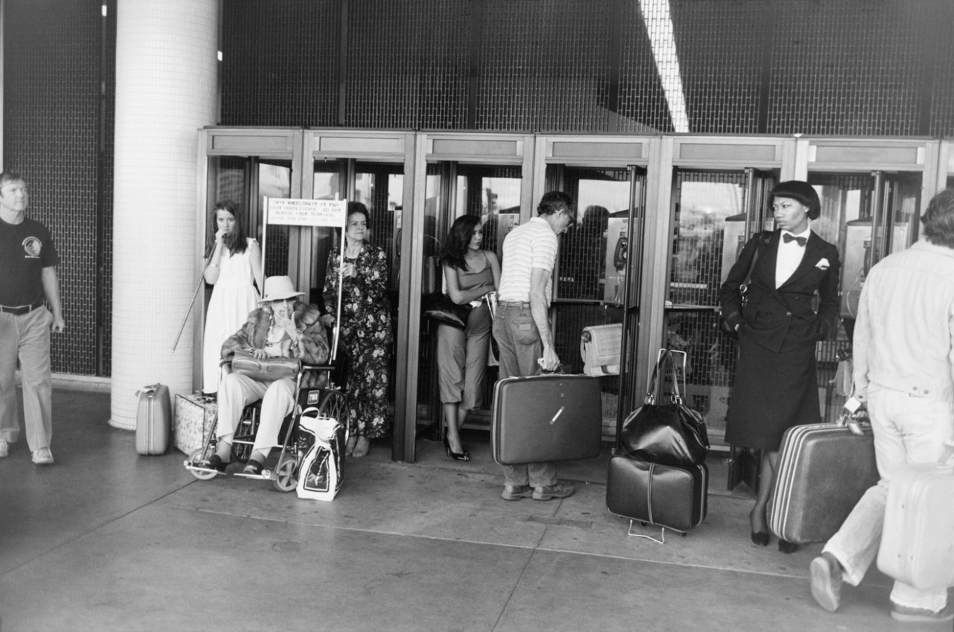 Black-and-white photograph of travelers with their suitcases outside a set of glass doors