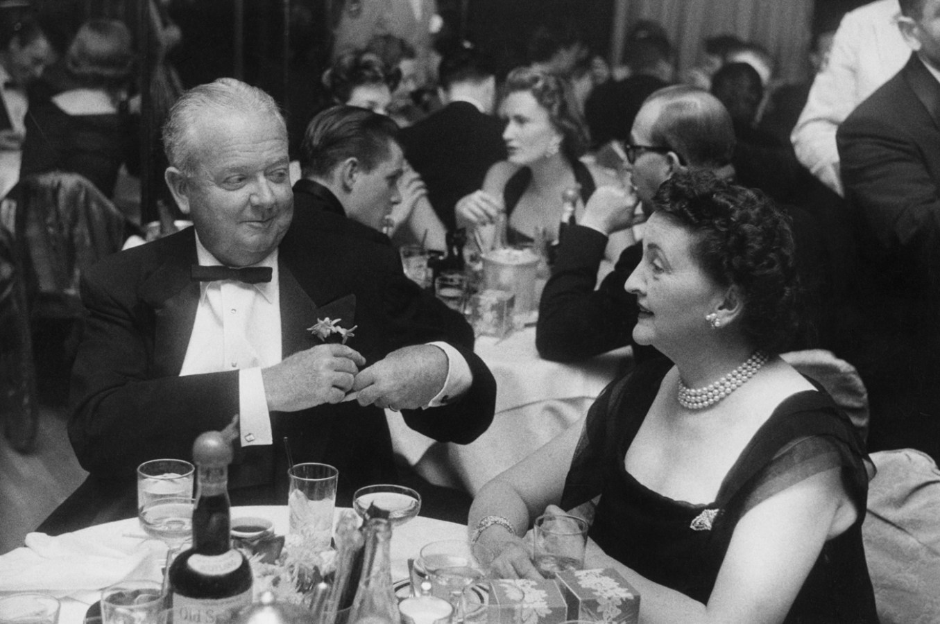 Black-and-white photograph of an older man and woman in formal dress conversing at a round table