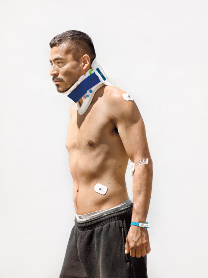 Color photographic portrait of a shirtless man in a neck brace standing in front of a blank white wall