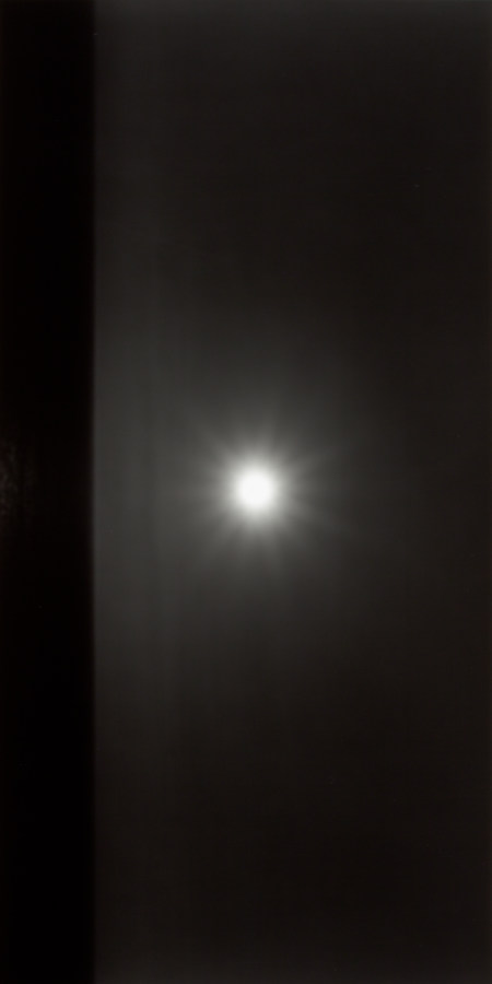 Black-and-white vertical photograph of a horizontal black seascape with a white round sun in a gray sky