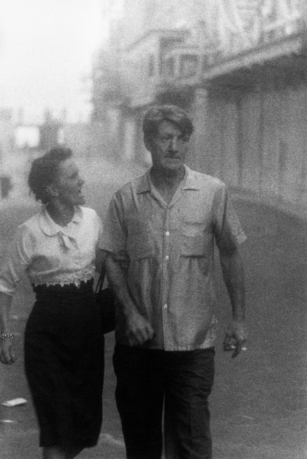 Black and white photograph of a couple walking down a boardwalk with the woman in mid-sentence