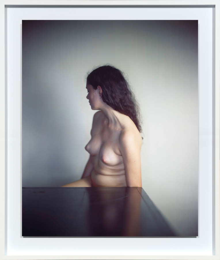 Color photographic portrait of a nude young woman seated at a barle table with her head turned back towards the gray wall