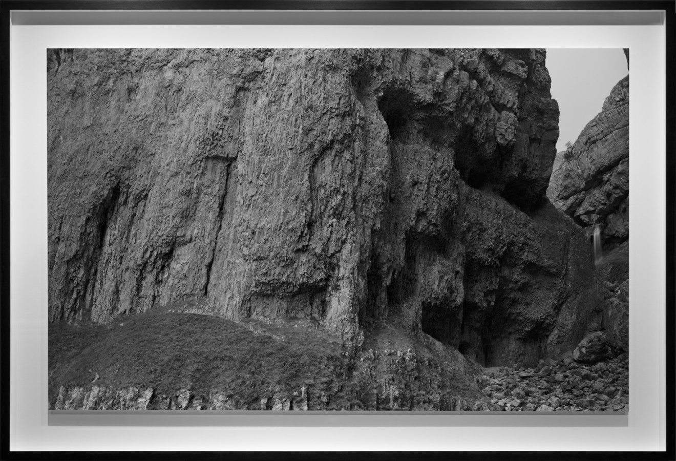 Black-and-white photograph of the stony base of a rough bare cliff face