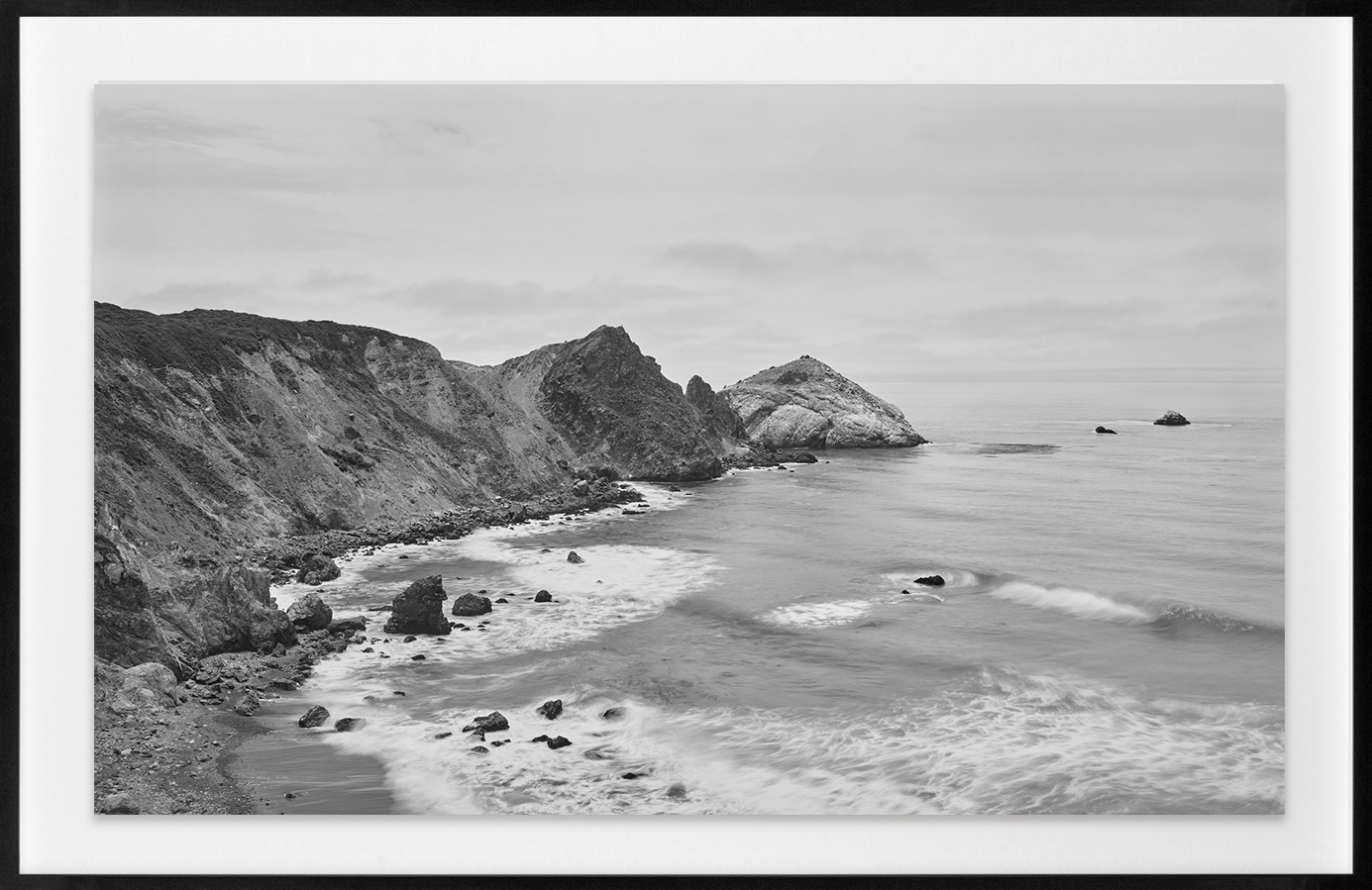 Black-and-white photograph of a cape curving around a bay with waves crashing on a rocky shore