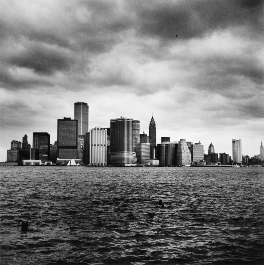 Black-and-white photograph of a city skyline from a river under a cloudy sky