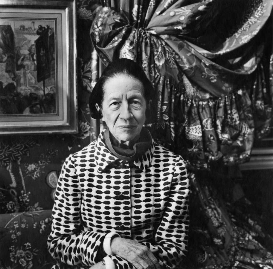 Black-and-white photograph of an older woman in a black-and-white patterned jacket standing in front of pinned-back floral curtains