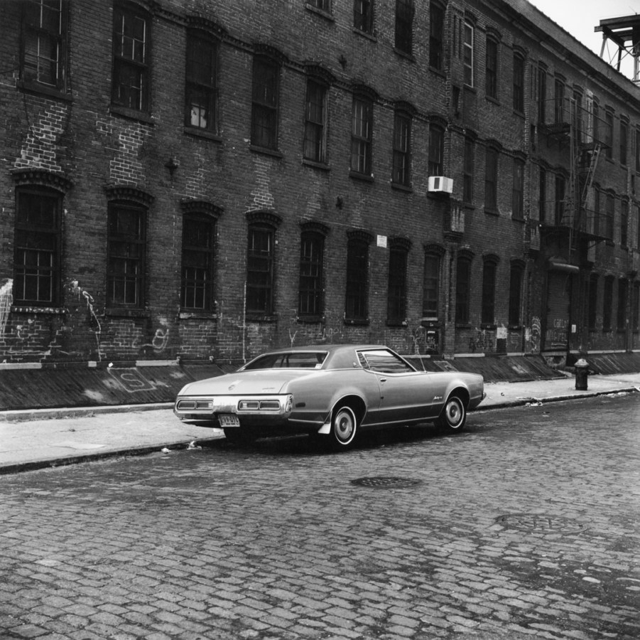 Black-and-white photograph of a car parked on a cobbled street next to a brick building