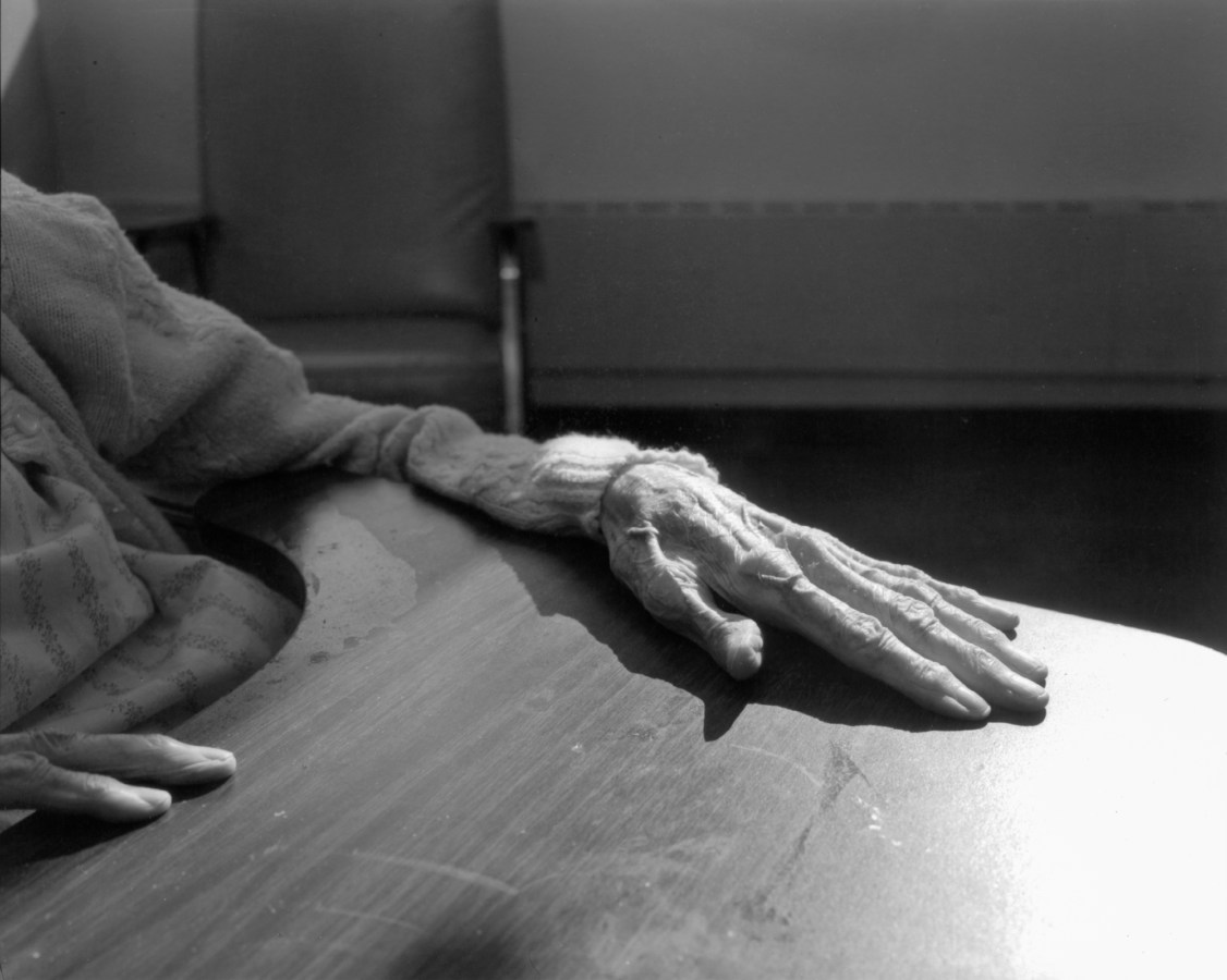 Black-and-white photograph of the hand of an elderly person resting on a tray table