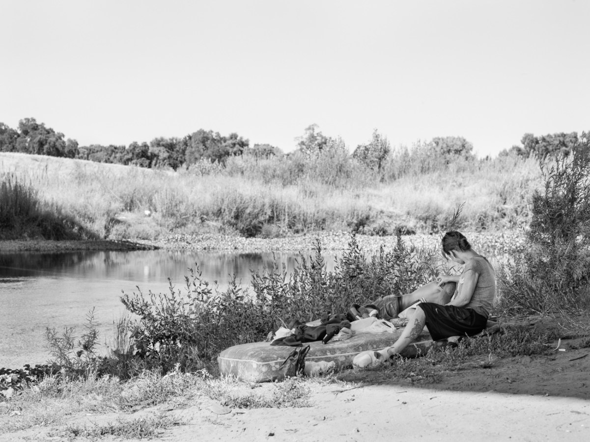 Black-and-white photograph of two people reclining on a bare mattress on a riverbank in the shade