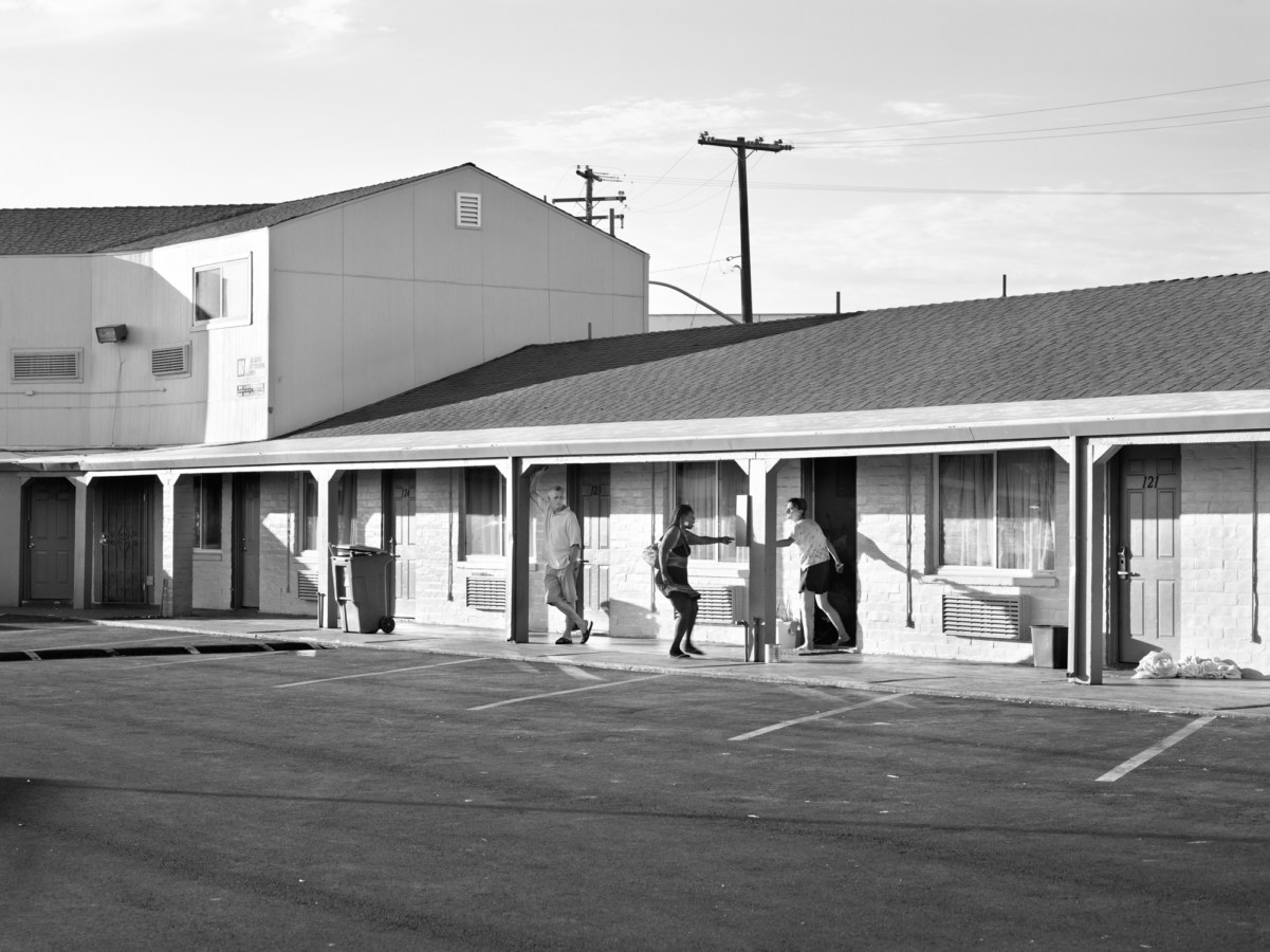 Black-and-white photograph of a a woman pointing at another coming out of a room in a motel parking lot with a man leaning on a pillar