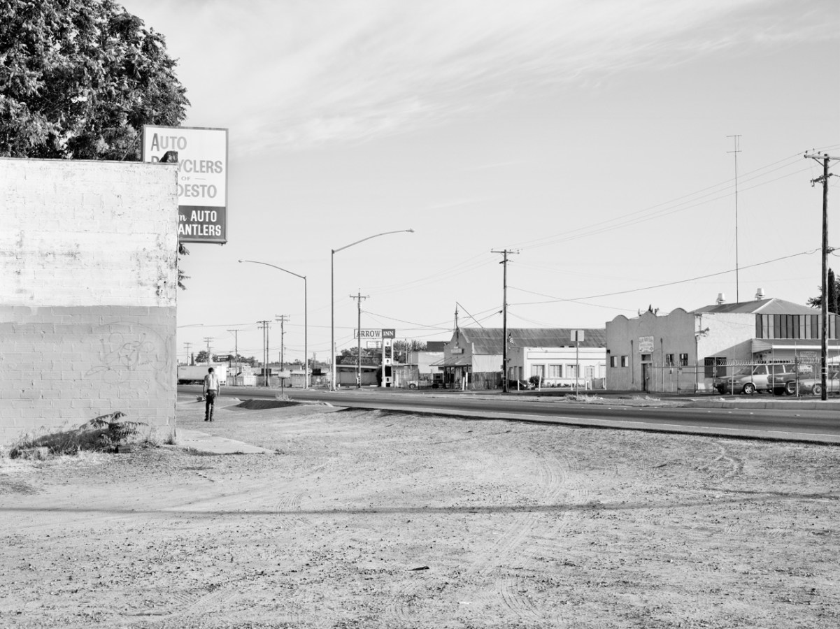 Black-and-white photograph of a dirt lot on a road with low buildings and a person walking away in the distance