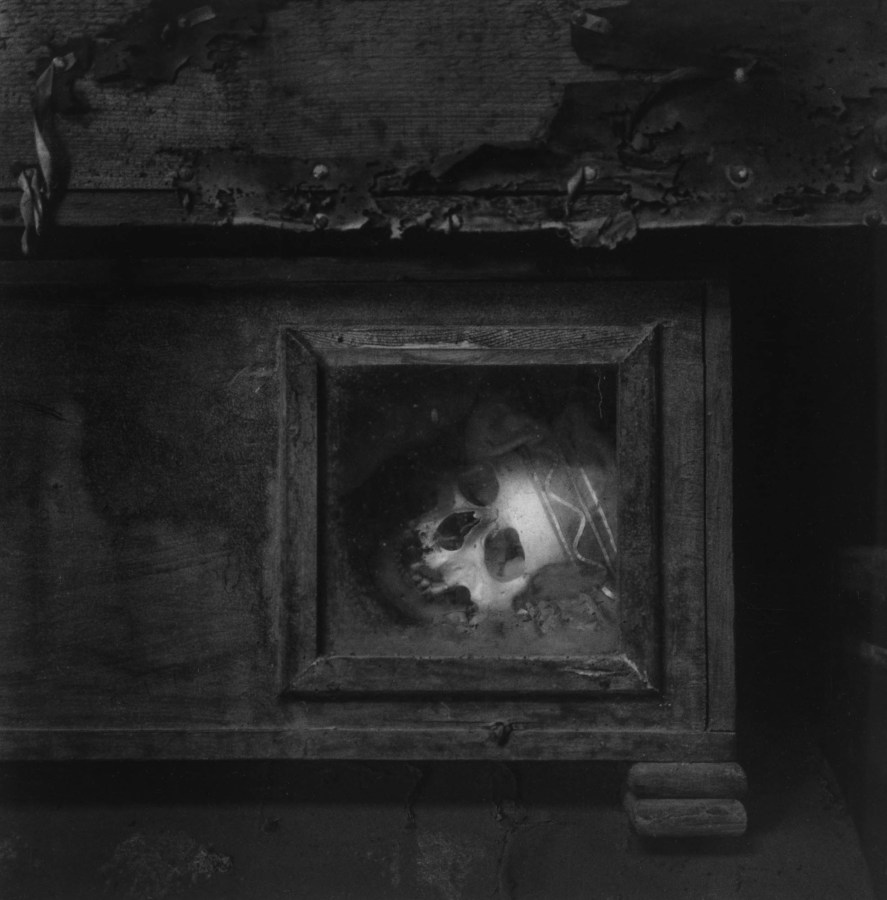 Black-and-white photograph of a human skull peering out of a glass-fronted wooden box