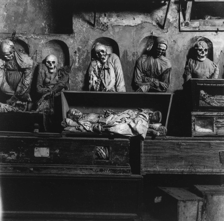 Black-and-white photograph of preserved shrouded bodies standing above wooden boxes containing children's preserved bodies