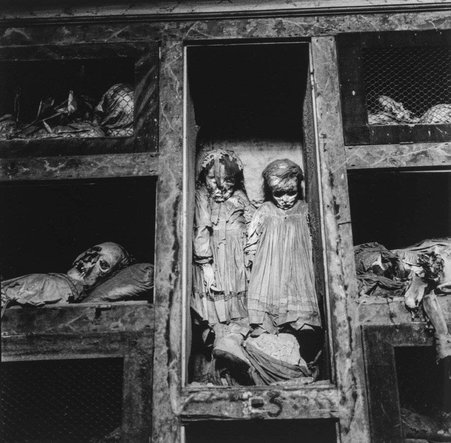 Black-and-white photograph of two preserved children's bodies propped up in a wooden compartment amid other bodies