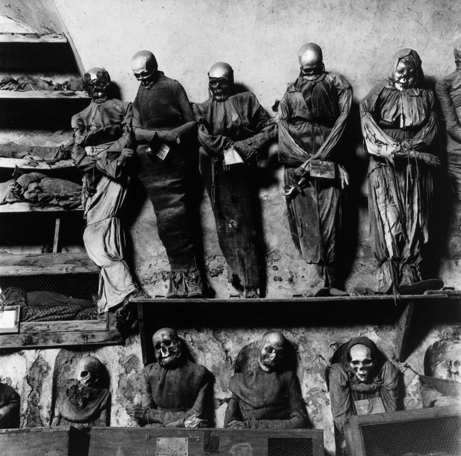 Black-and-white photograph of preserved skeletons dressed in shrouds and gowns in two rows