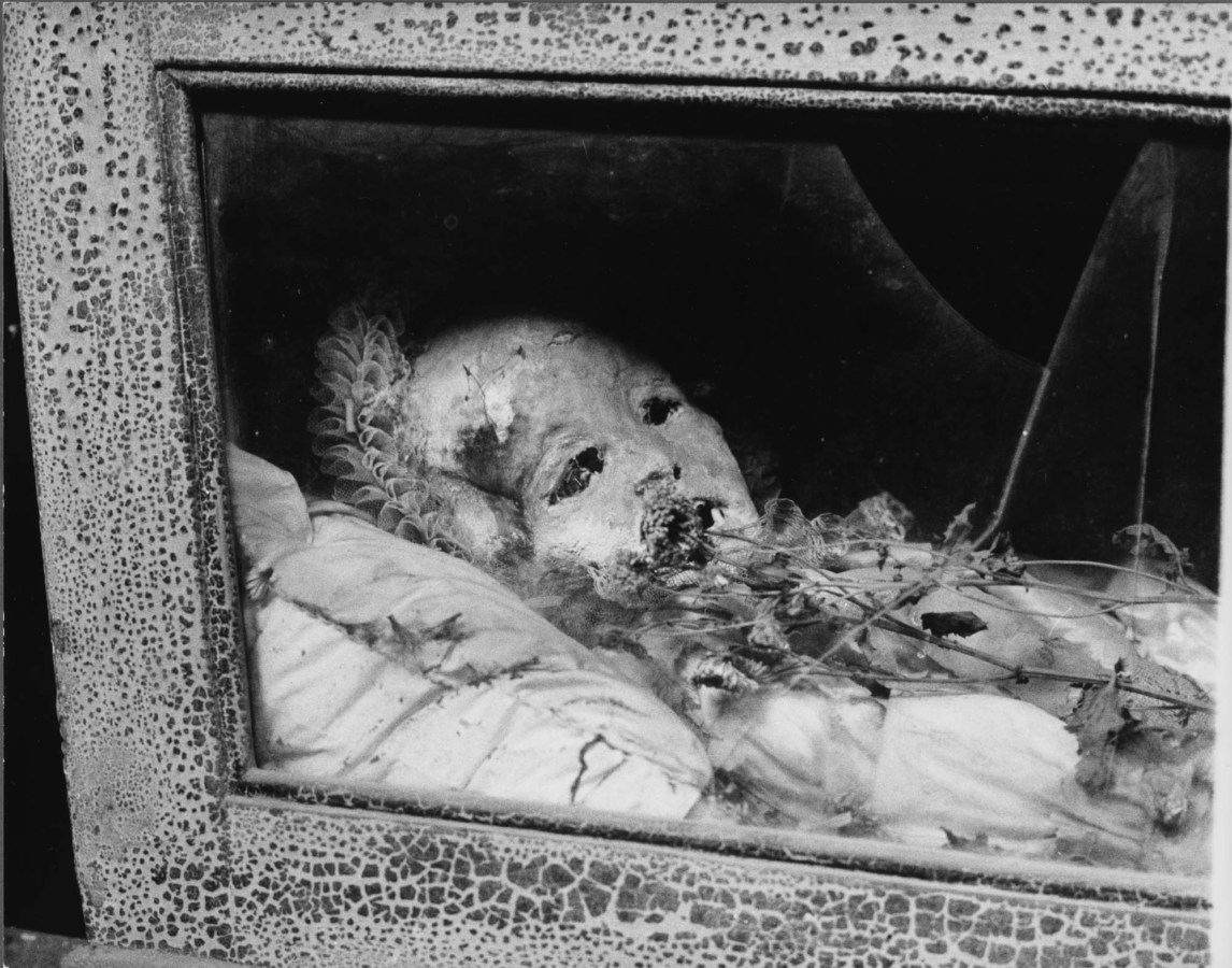 Black-and-white photograph of a preserved child's body lying in a glass-fronted wooden case with dried flowers