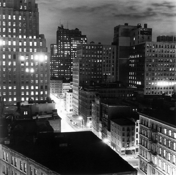 Black-and-white photograph of city buildings with lighted windows above a glowing street
