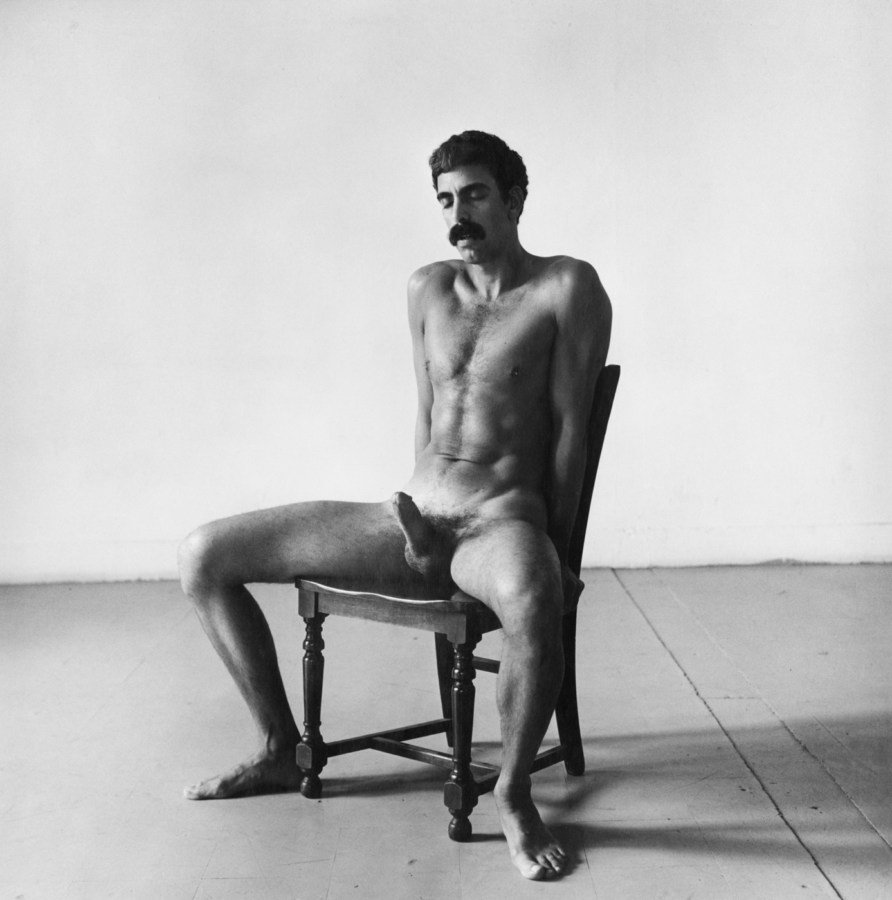 Black and white photograph of a seated nude man with an erection and his hands behind his back