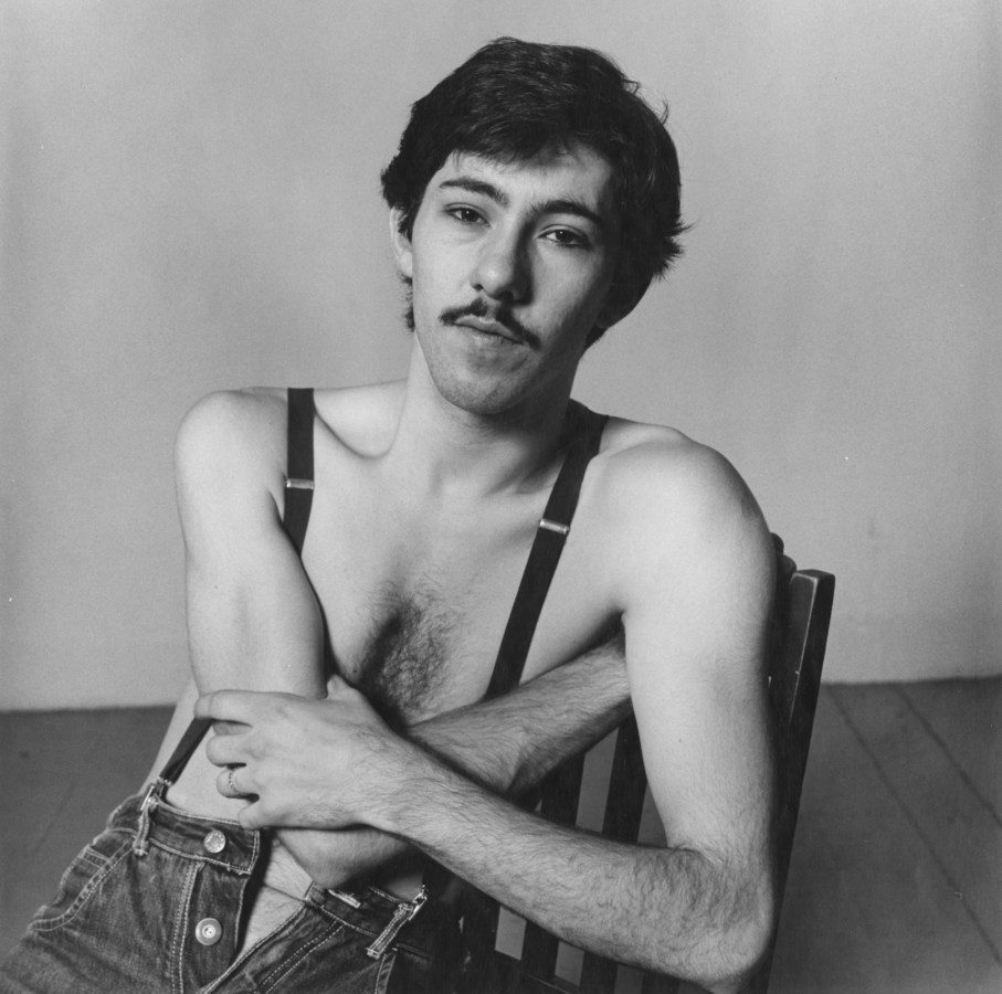Black-and-white photograph of a seated man with a mustache wearing only suspenders and jeans