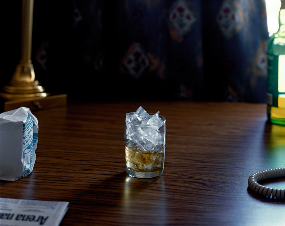 Color photograph of a glass of whiskey with ice on a dark wood desk