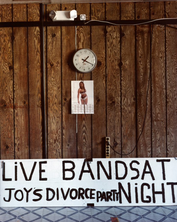 Color photograph of a wood paneled wall with a clock, picture of a woman in a bikini and hand-painted sign reading LIVE BAND JOY'S DIVORCE PARTY SAT NIGHT