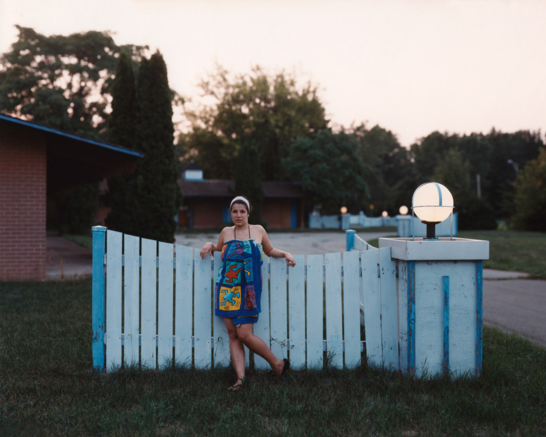 Color photograph of a woman wrapped in brightly patterned towel leaning against a blue fence
