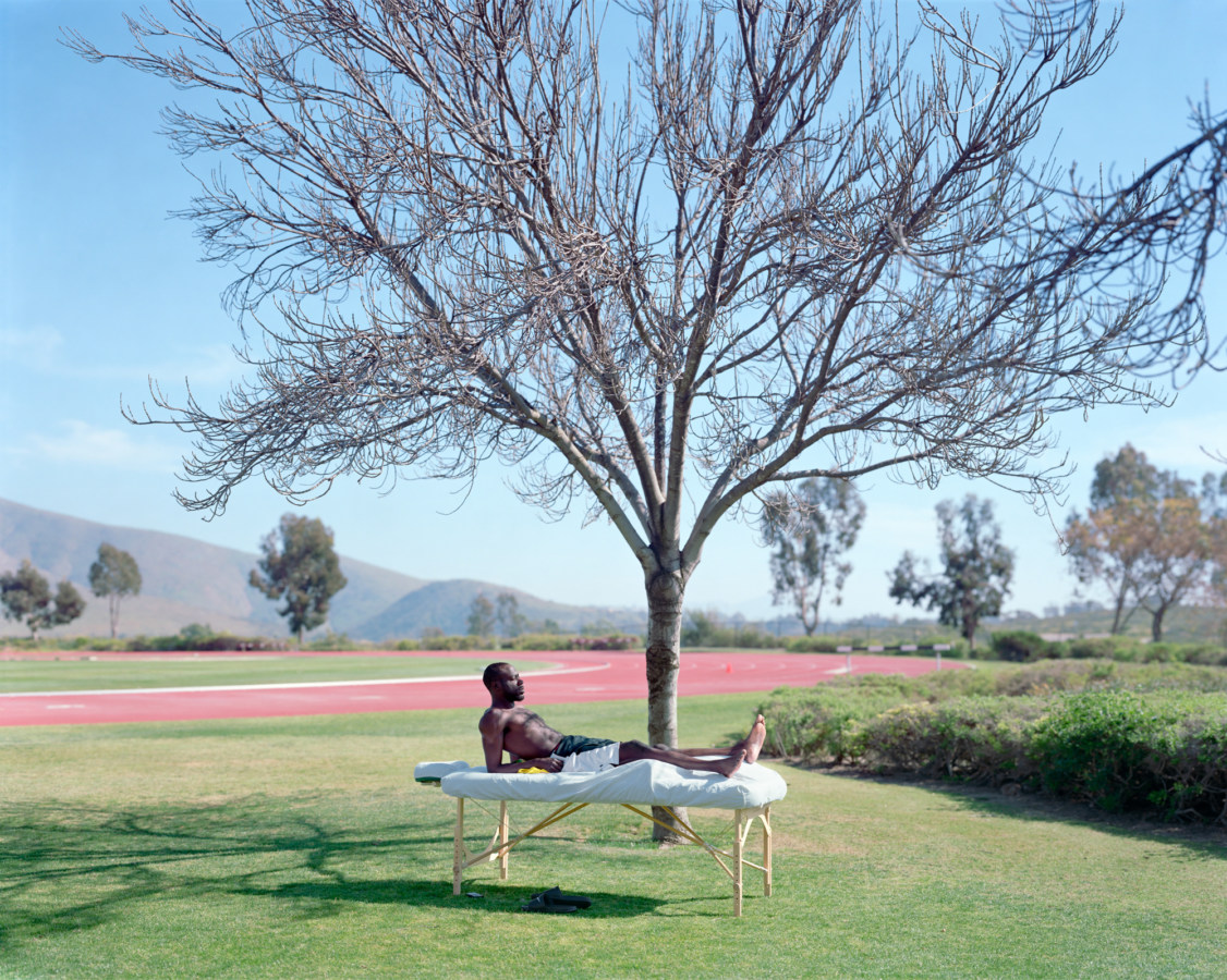 Color photograph of a man reclining on a massage table under a bare tree on a green lawn next to a running track