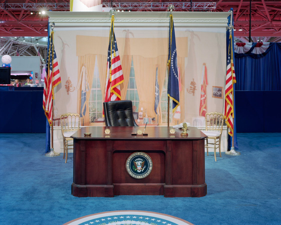 Color photograph of an empty set of the Oval Office with desk and painted backdrop in a convention hall setting