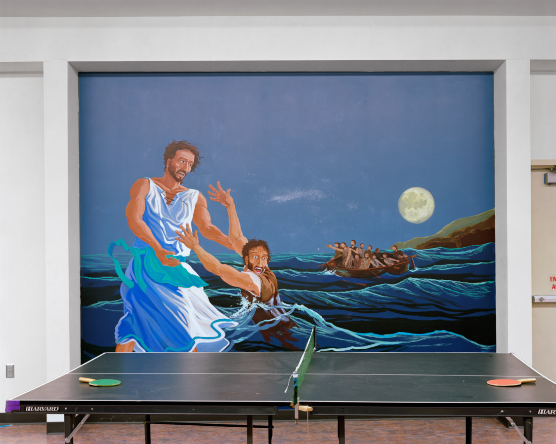 Color photograph of a ping pong table in front of a mural of a man reaching out of the sea to another dressed in white walking on water