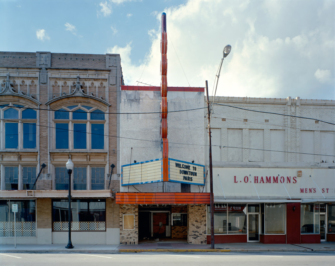 Color photograph of a movie theater with an oversized neon sign wedged between two other empty storefronts