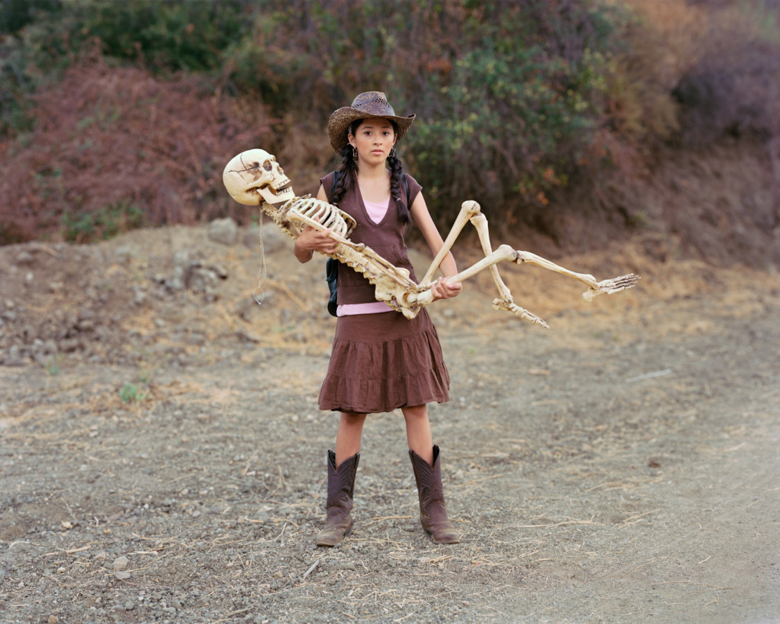Color photograph of a girl in a cowboy hat and boots cradling a life-size plastic skeleton