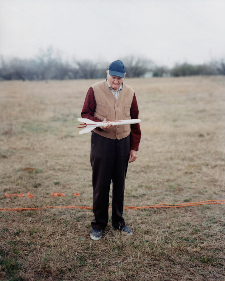 Color photograph of an elderly man standing on a brown lawn looking down at a model rocket in his hand