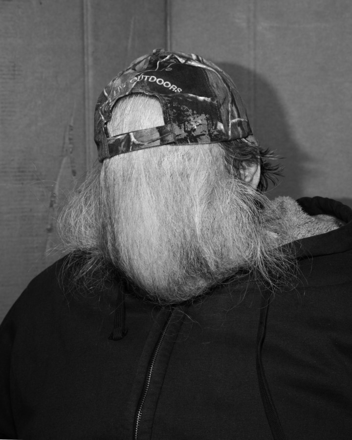 Black and white photograph of a person using their beard hair to cover their face tucked into their hat