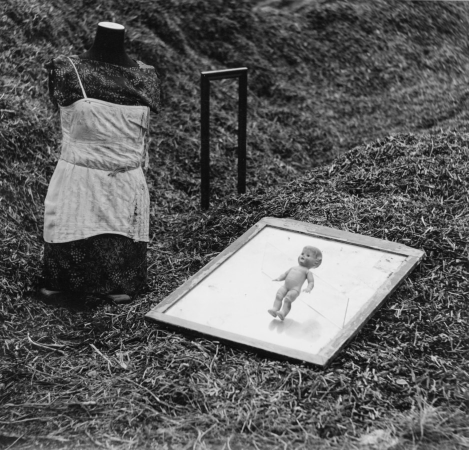 Black-and-white photograph of a headless mannequin positioned on the ground next to a small doll hovering above a tray