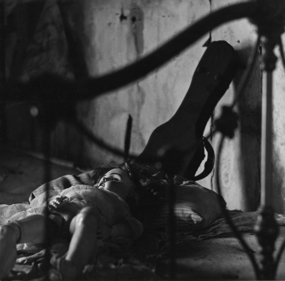 Black-and-white photograph of eyeless doll lying on a bed through the metal bedframe