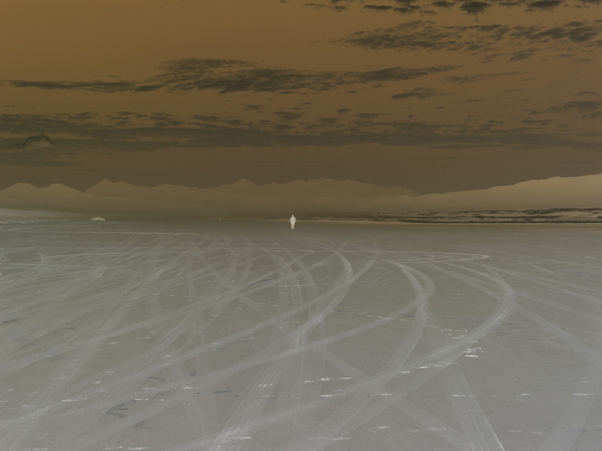 Inverted color photograph of a person standing on a sandy beach with a mountain range on the horizon