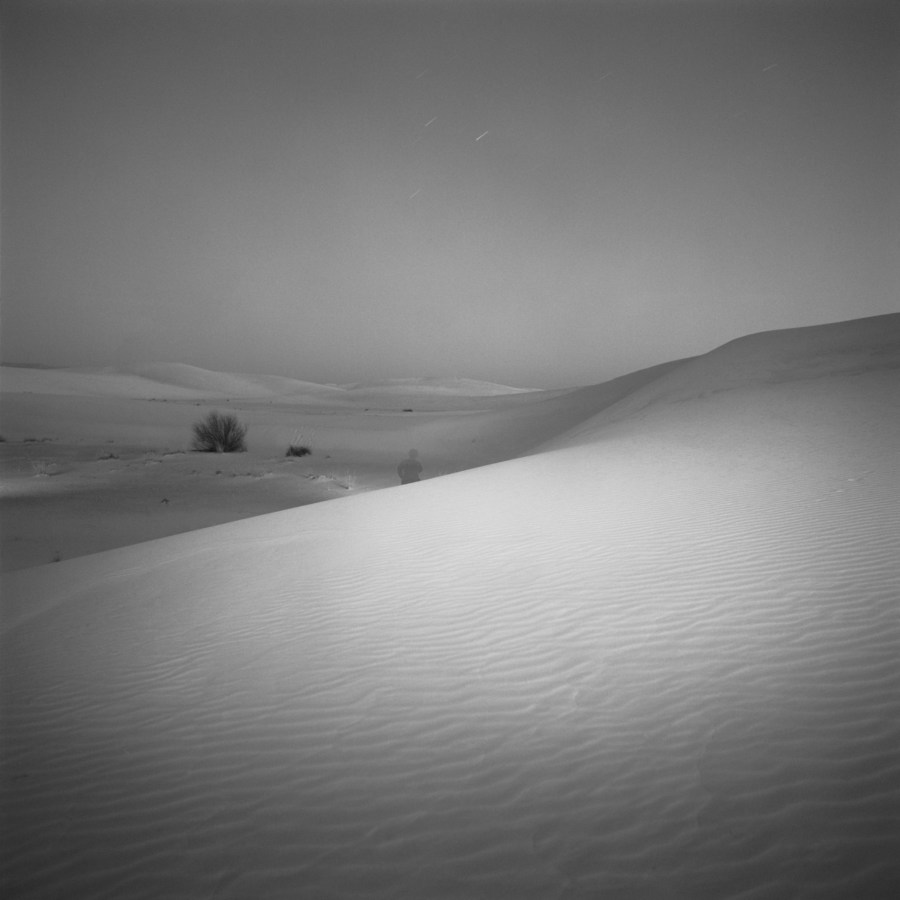 Black-and-white square photograph at night of a single figure amid a valley of sand dunes