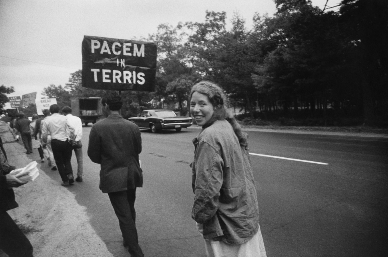 Black-and-white photograph of a woman smiling turning around mid-step following a man with a sign reading PACEM IN TERRIS in a line of people holding signs