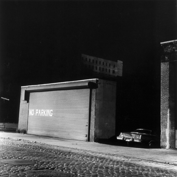 Black-and-white photograph of a shuttered garage door painted with NO PARKING and single car parked in an adjacent driveway on a cobbled street