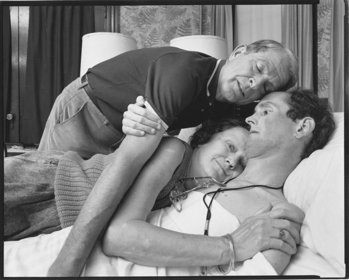 Black and white photograph of two people embracing a third individual who is lying down in bed