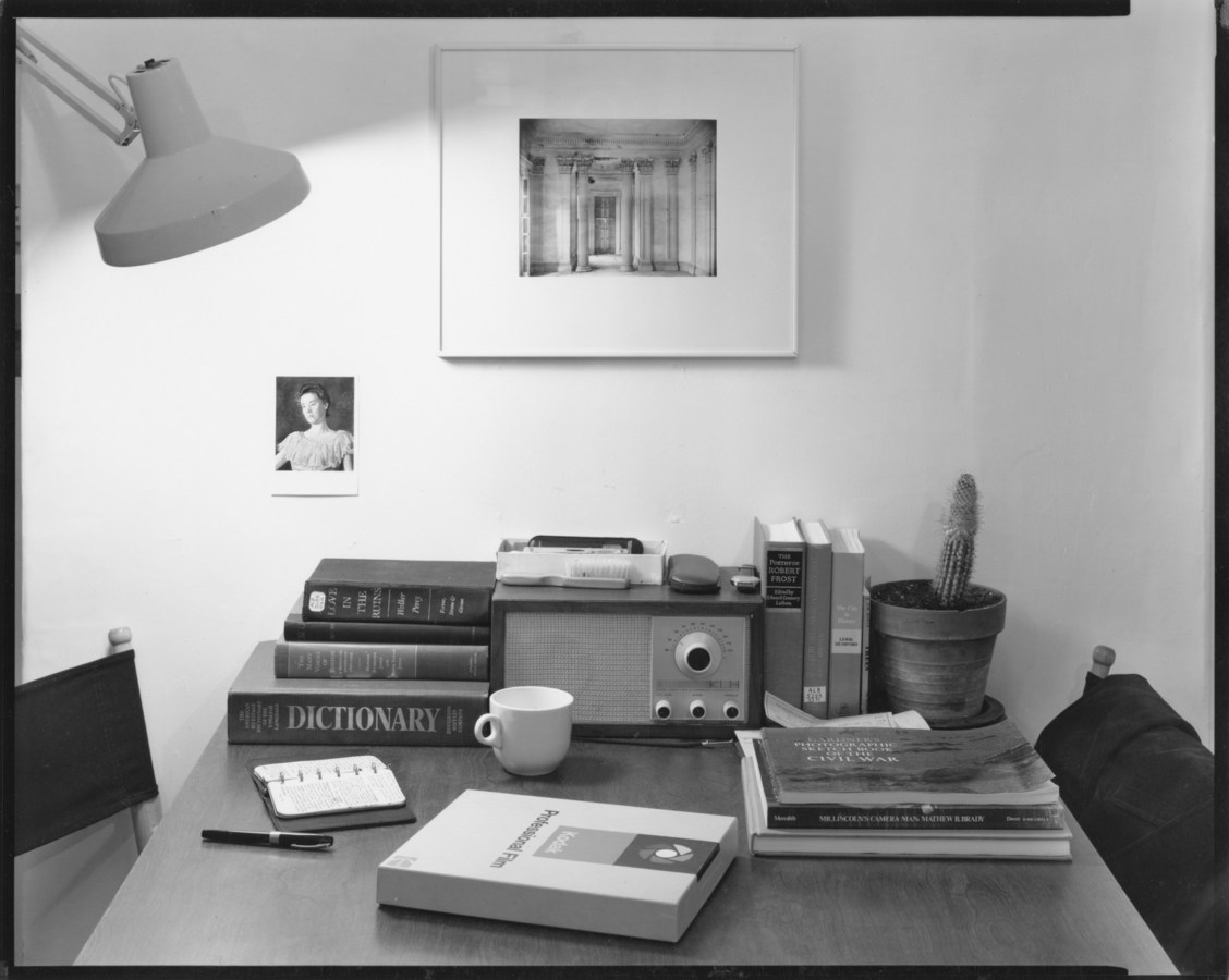 Black and white photograph of a desk with books, cactus, coffee mug, box of film, radio, and notebook illuminated by a standing lamp