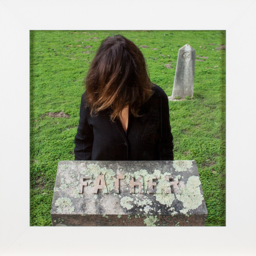 A framed square photograph of a woman in a graveyard, behind a tombstone reading "Father," with her hair in front of her faace