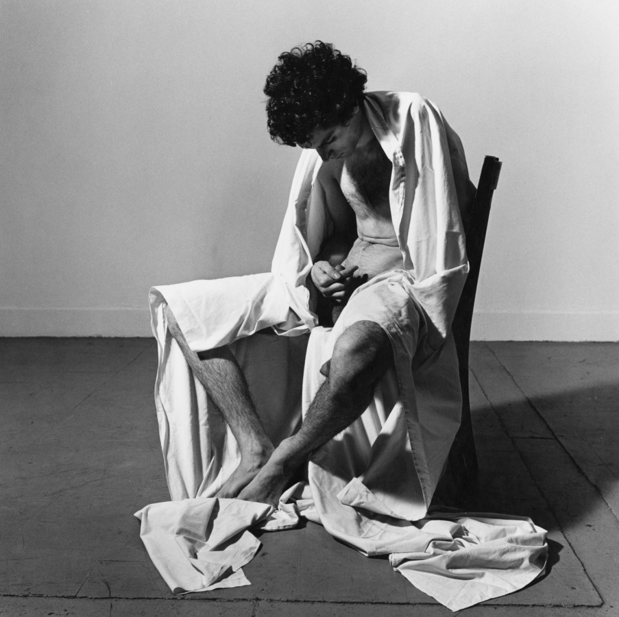 Black and white photograph of a nude man seated on a chair, draped in a sheet