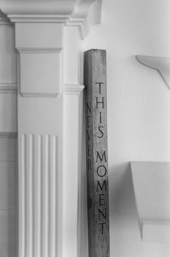 Black-and-white photograph of a column and an engraved wooden staff which reads "This Moment"