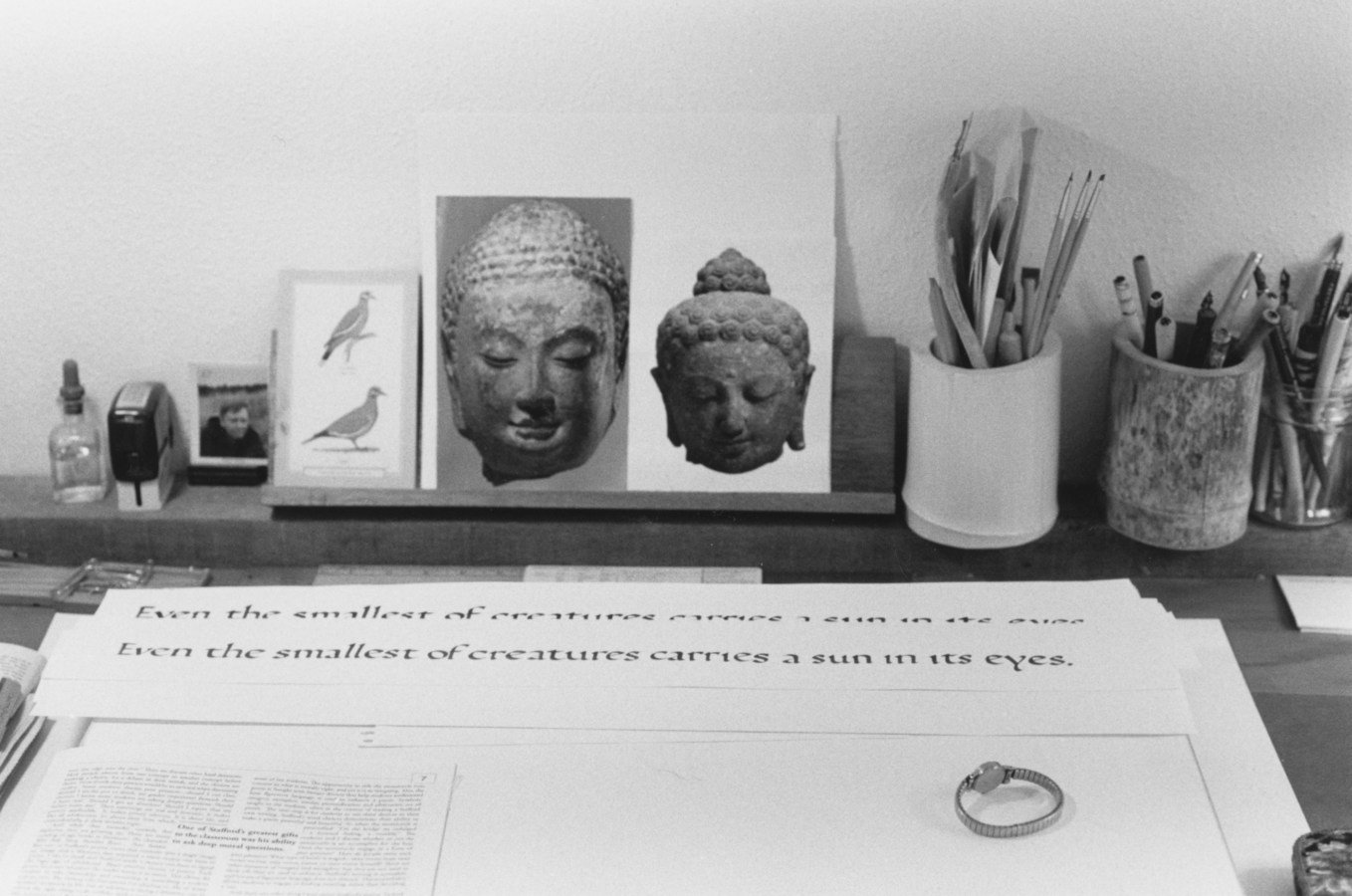 Black-and-white photograph of a tabletop with calligraphy writing, brushes, and postcards of Buddha head sculptures.