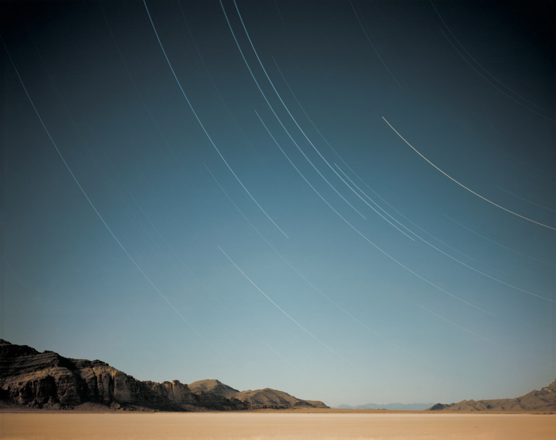Color photograph of a beach with a mountain range on the horizon under a sky filled with long-exposure curved star-trails