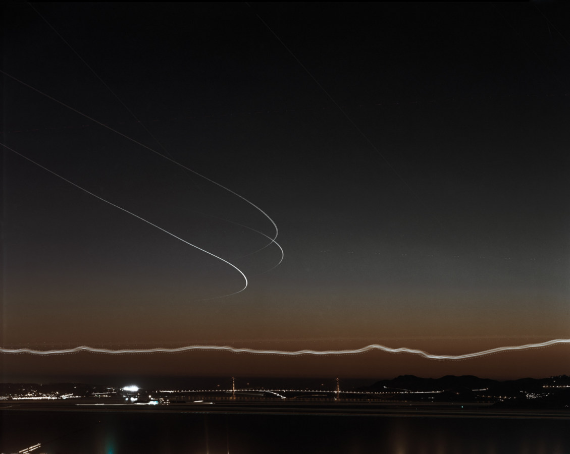 Long-exposure color photograph of the distant Golden Gate Bridge just after sunset under light trails of airplanes